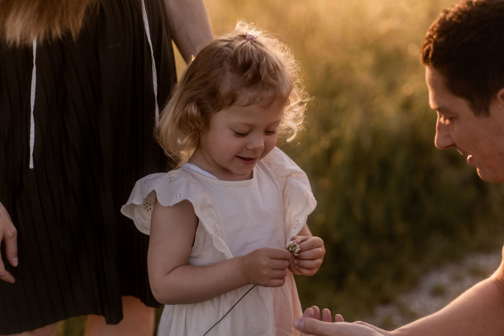 Kleines Mädchen im weißen Kleid hält Blume in der Hand, Vater sitzt knied vor ihr Mutter steht dahinter und genießen das Familienshooting von PIC@MO bei Sonnenuntergang