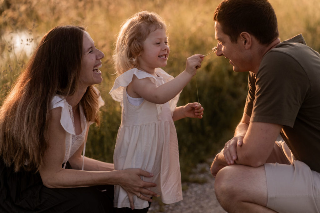Kleines Mädchen im weißen Kleid hält Blume in der Hand und kitzelt die Nase vom Papa. Mutter steht daneben und lacht. Im Hintergrund geht die Sonne unter