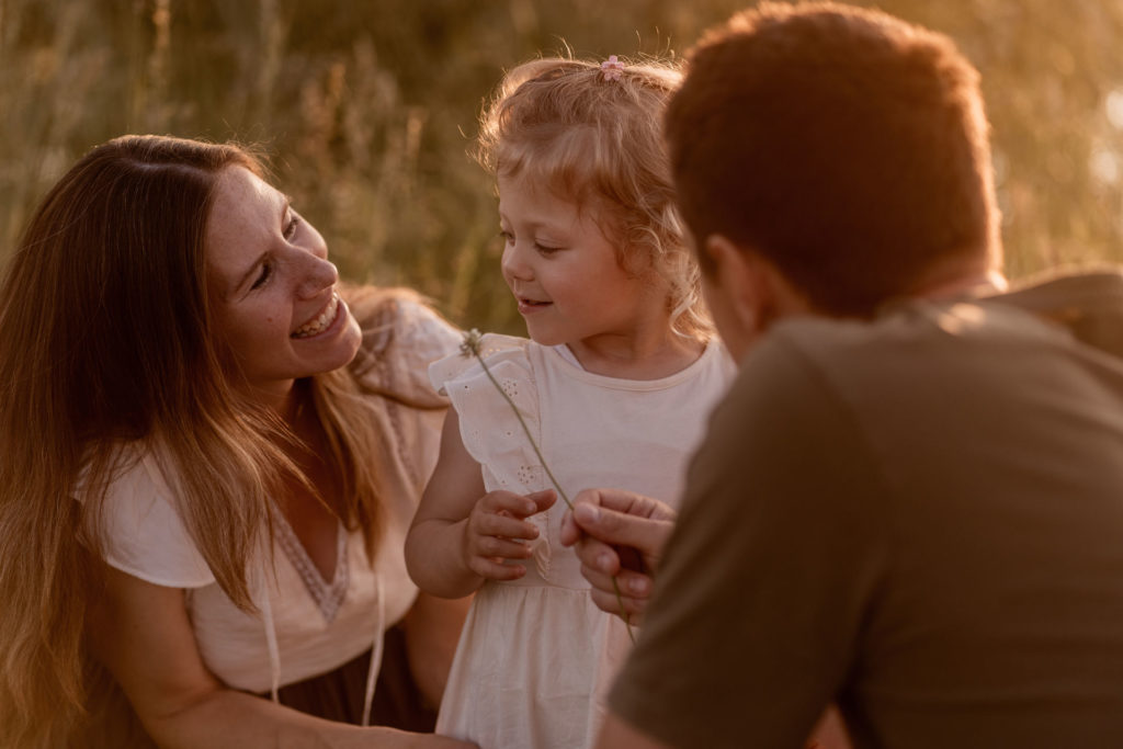 Familie in einer Wiese bei einem Familienshooting von PIC@MO mit Mona von Malm: Mutter lacht kleines Mädchen im weißen Kleid an. Vater schaut beide an