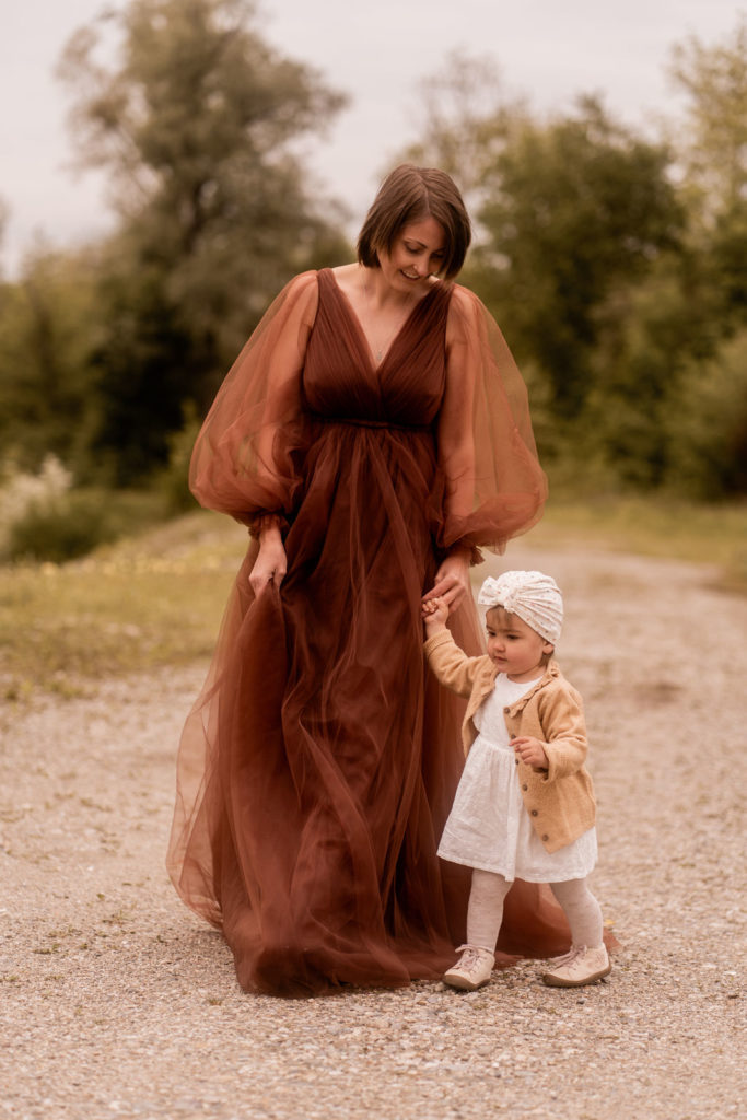 Mutter in einem braunen Kleid mit Kleinkind in der Hand spaziert auf einem Feldweg bei einem Mummy-and-me-Shooting von PIC@MO mit Mona von Malm
