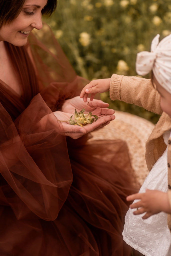 Mama und Kleinkind sitzen auf Wiese. Kleines Mädchen legt Blumen in die Hände der Mutter.