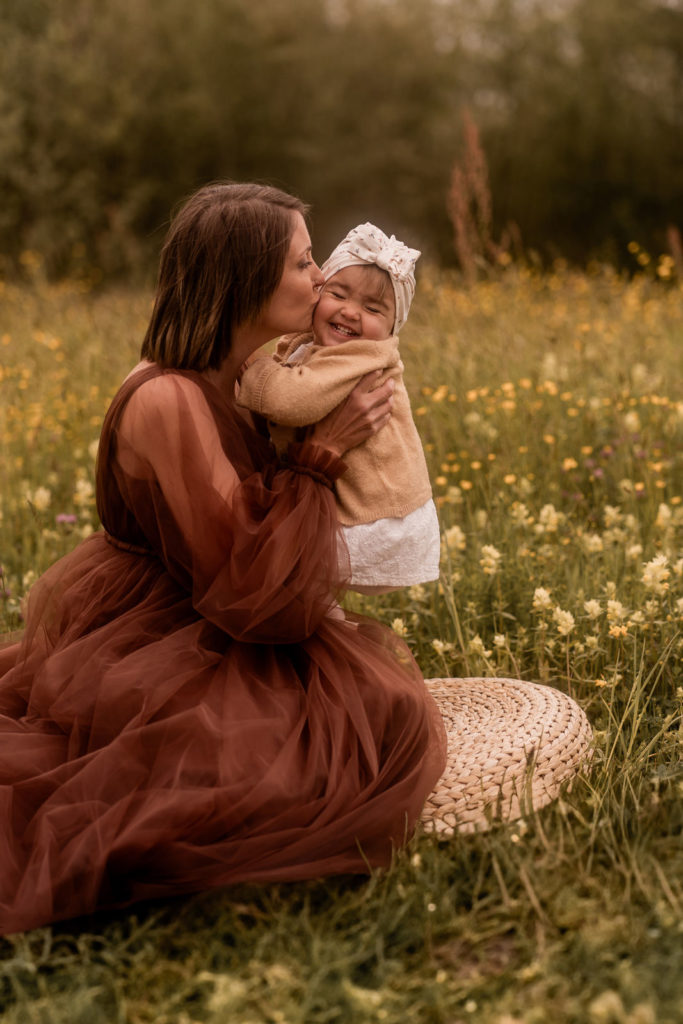 Mutter im braunen Kleid sitzt auf einer Wiese und hält kleines Mädchen hoch und küsst es auf die Wange