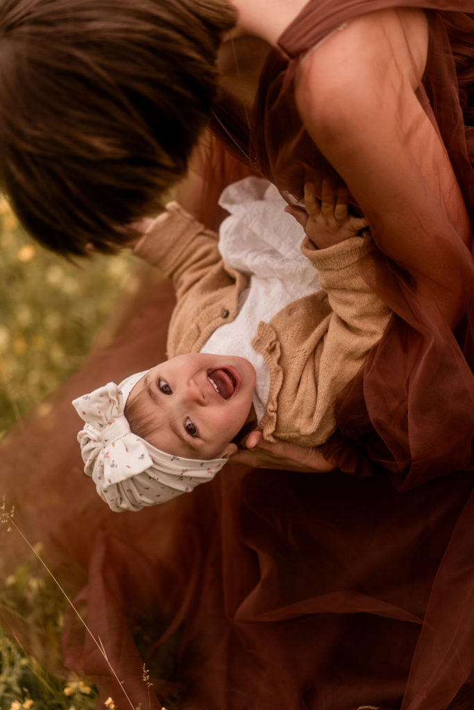 Mutter sitzt auf einer Wiese und hält Kind im Arm, dass in die Kamera lacht bei einem Mummy-and-me-Shooting von PIC@MO mit Mona von Malm